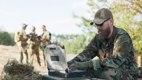 grupo de soldados que esperan para entrar en acción en el campo vestidos, preparados con equipo y armas esperando en la distancia para el comandante, que se sienta en la computadora y envía datos a la base