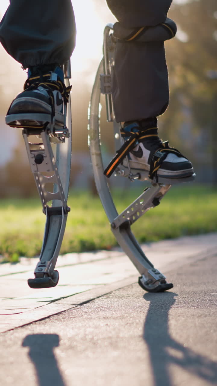 Person on spring stilts walking on paved path in sunny park, captured from low angle focusing on legs in dark pants. Scene highlights dynamic movement, vibrant energy, and natural outdoor
