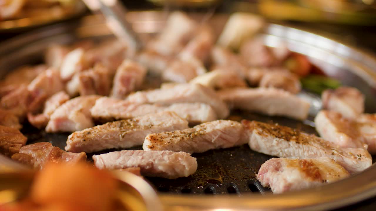 Close-up of pork grilling on hotplate, tongs turning meat, warm lighting, shallow depth of field
