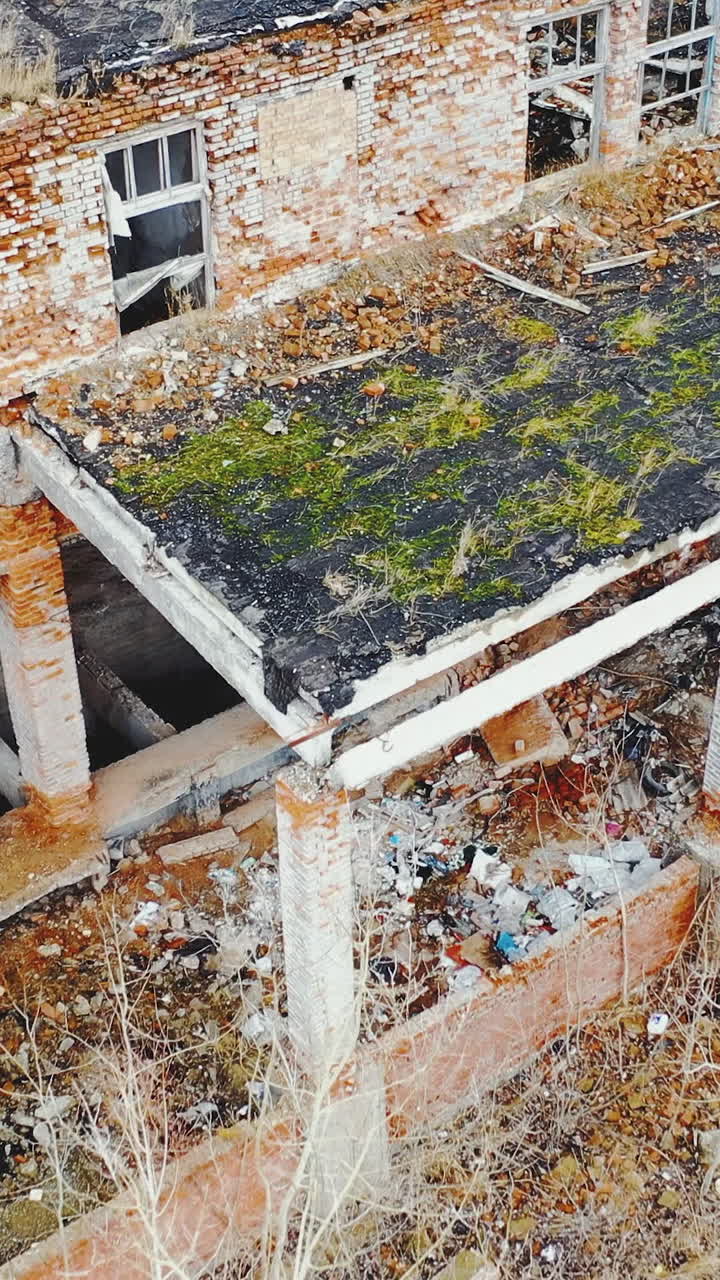 Ruined brick buildings with collapsed roofs. Broken dirt road with heaps of garbage. Moss and grass on the ruins. Aerial view. Vertical video