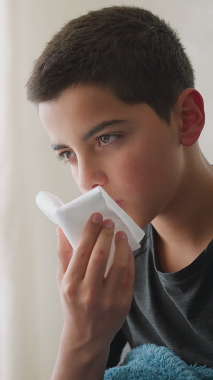 Exhausted young boy sneezing into white napkin covered with blanket, using tissue to wipe his nose while resting on the couch near the window, showing signs of tiredness