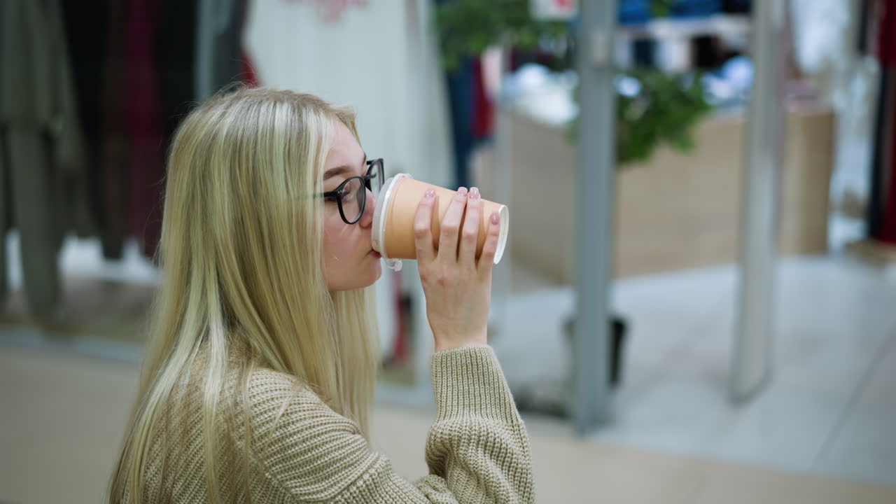 dama bebiendo café de la taza mientras usa tela de punto en un centro comercial con fondo borroso mostrando ropa de una tienda, creando una experiencia de compras acogedora con un ambiente relajado