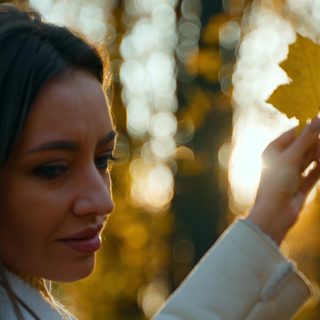 Donna con capelli lunghi e giacca bianca che tiene in mano una foglia gialla d'acero, girandola. Luce solare nel parco autunnale sullo sfondo, in modalità sfocata