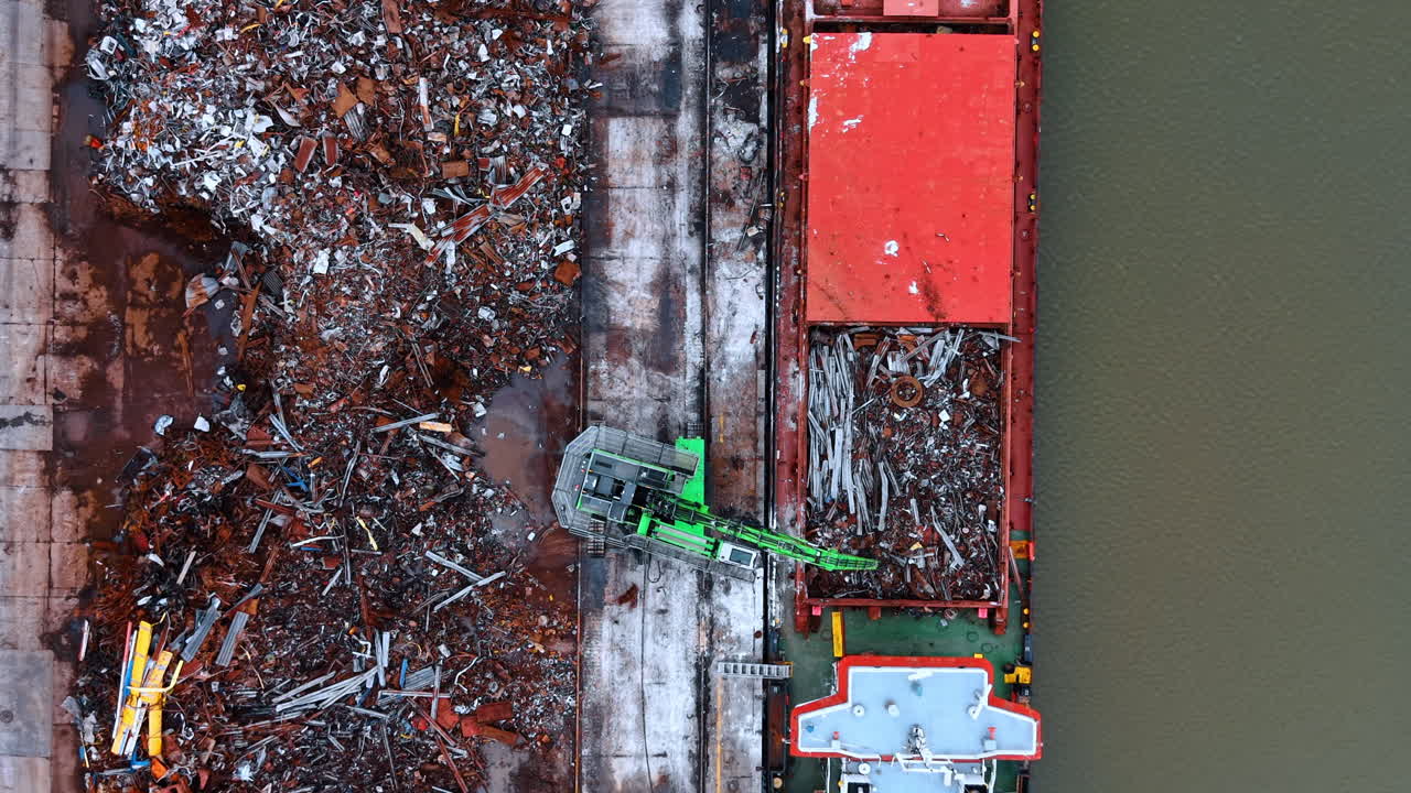 Process of loading metal waste into a bulk carrier hold. Top view of a green hydraulic manipulator moving scrap metal from the pier into the ship's cargo hold