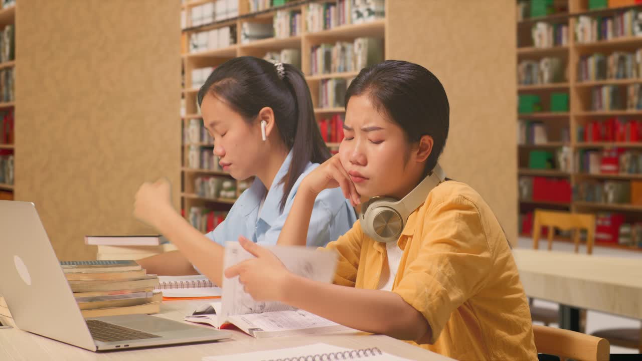 헤드폰을 착용한 아시아 여성 학생들은 도서관에서 노트북과 함께 테이블에 앉아서 책을 읽는 것에 지루합니다.