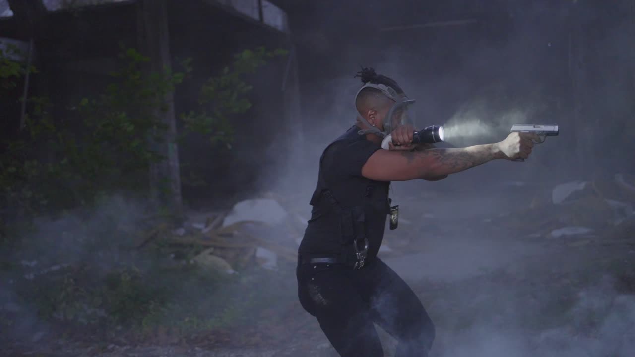 연기가 나는 건물에 있는 흑인 경찰이 용의자를 찾기 위해 주변을 감시하고 있습니다.