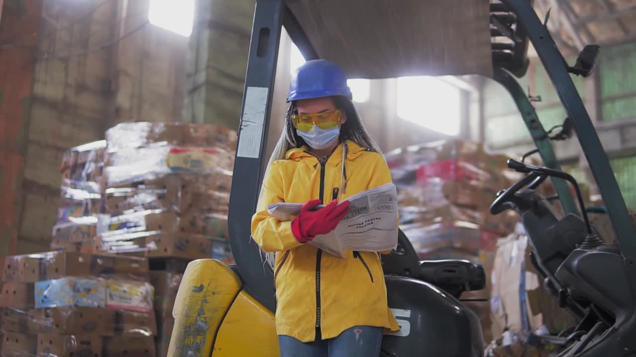 Female worker in hard hat and yellow jacket standing, leaning on warehouse electric car and reading work journal. Huge stocks of pressed carton. Low angle view