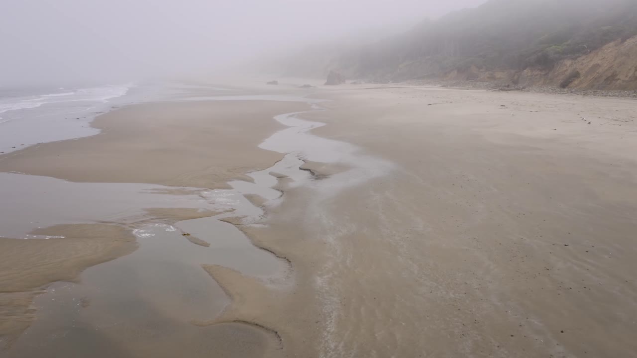 Low tide at Cannon Beach, Oregon Coast in a rainy, moody and foggy day. Dolly in drone proximity aerial over gold sand tidal flats
