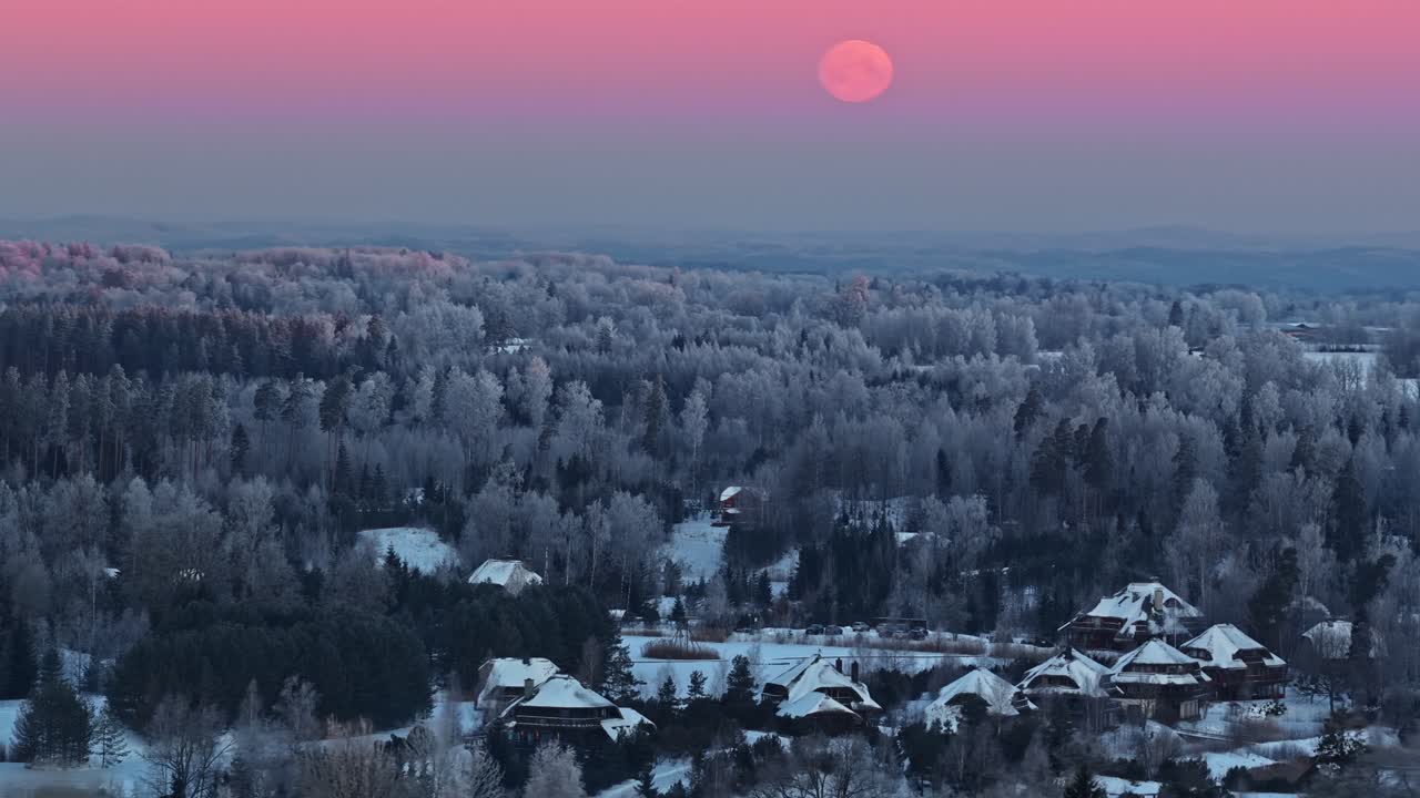 Fuldmåne over en frostklar skov og sneklædt landsby ved vintertusmørke, med lyserød pastelfarvet himmel og roligt luftlandskab med plads til tekst.