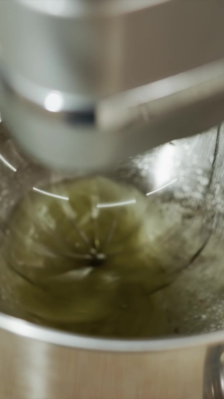 Primer plano de una batidora de pie mezclando masa verde. La mezcla se espesa a medida que es continuamente mezclada por las varillas giratorias. Una pequeña cantidad de polvo blanco aparece cerca de la base sobre superficies de madera.