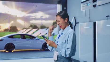 Side View Of Asian Female Professional Worker With Her Smartphone With Home Energy Storage System In a Modern Garage, She Raises Her Fist Up With Screaming Goal After Check On The Smartphone