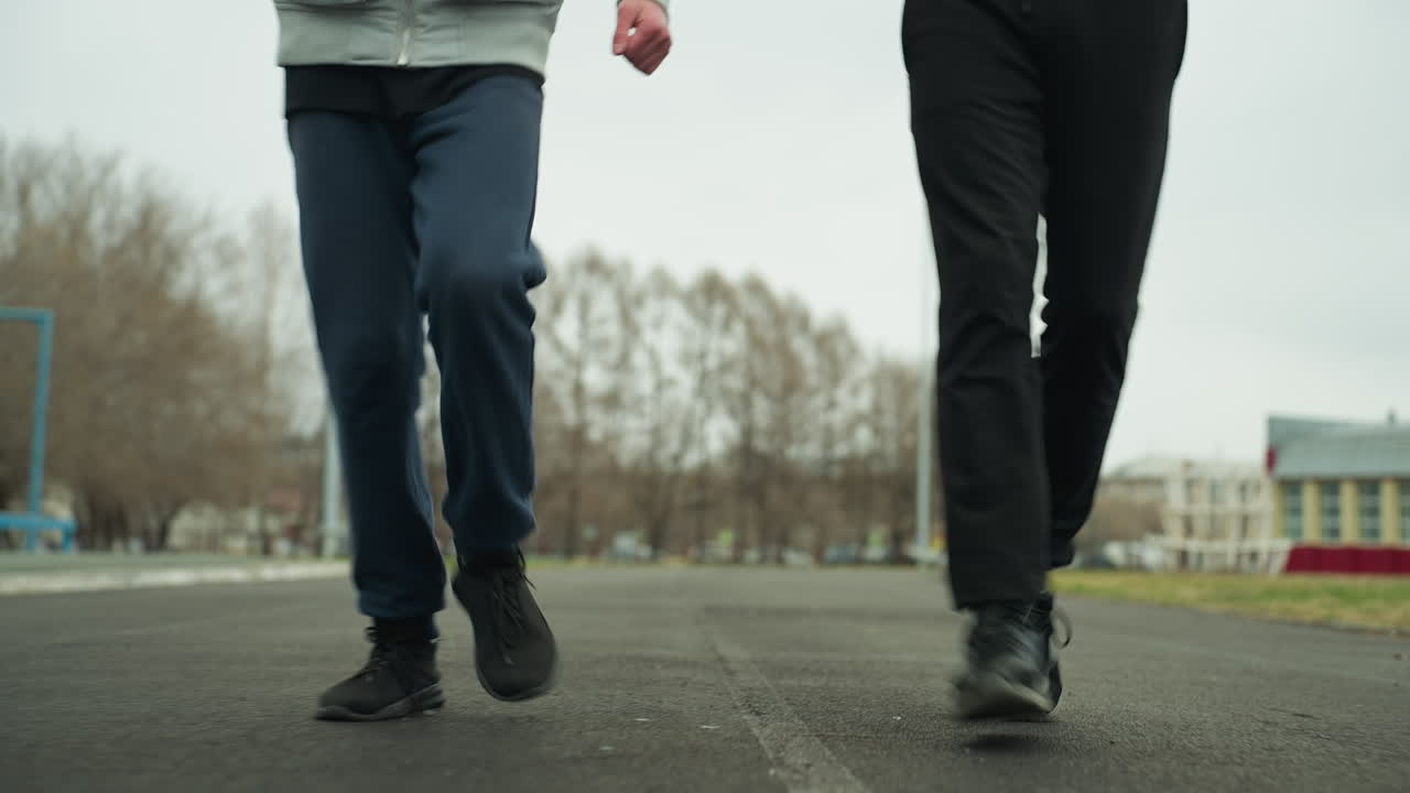 Close-up leg view of two people jogging on a track in a stadium, the background shows blurred light poles and parked cars with bare trees