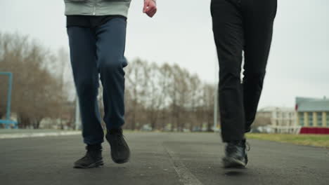 close-up leg view of two people jogging on a track in a stadium, the background shows blurred light poles and parked cars with bare trees