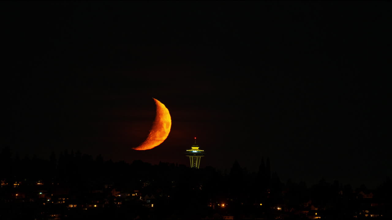 Striking timelapse of a glowing crescent supermoon setting over Seattle, with the Space Needle illuminated below, creating a captivating contrast against the dark sky.