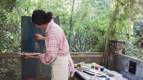 Biracial-female-artist-in-apron-painting-on-canvas-in-sunny-garden,-slow-motion