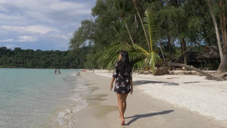 Asian-Model-in-a-short-black-flower-dress-walking-on-a-tropical-beach-on-the-shoreline,-Slow-Motion