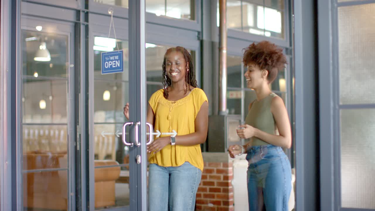 Donne afroamericane con maglie gialle e verdi che girano il cartello 'chiuso' e tirano le porte a vetri per aprire il negozio.