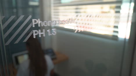 Transparent-phone-booth-in-a-coworking-office-space-showing-a-women-working-at-her-laptop