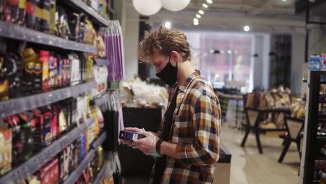 cliente en el supermercado durante el tiempo de la pandemia. hombre leyendo la etiqueta en el paquete del café. estantes en el fondo. usando máscara protectora negra
