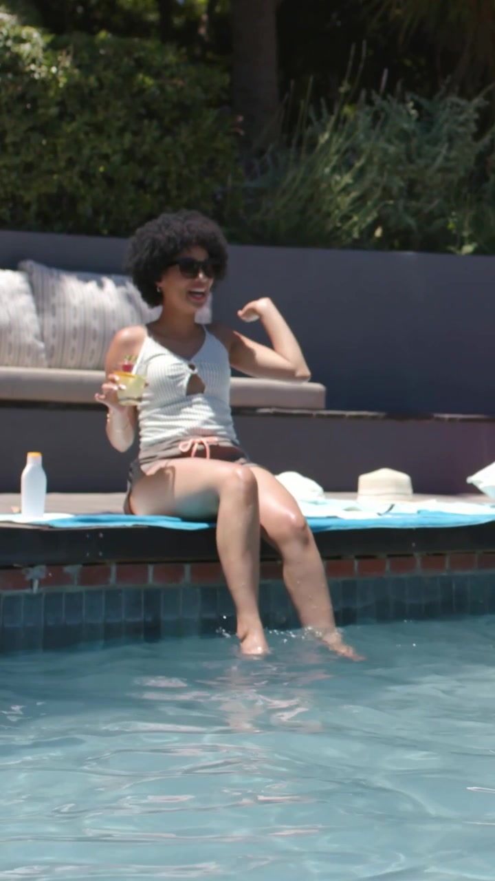 Vertikales Video: Lachende Frau in Shorts hält Becher mit Strohhalm am Poolrand, nachdem ein Freund sie bespritzt hat.