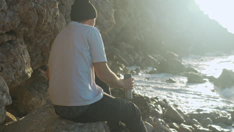 man enjoys himself on a rocky coast cliff shore drinks thermos coffee as sunrise sea waves crash