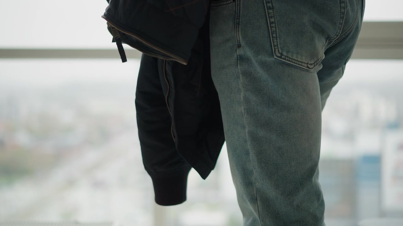 Close up of person lower body wearing light blue jeans and holding dark clothing in front of large window with blurred cityscape background. Jeans show visible texture details zipper fly