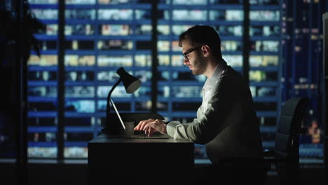 Hombre-De-Negocios-Enfocado-Trabajando-En-Una-Computadora-Portátil-Tomando-Notas-Sentado-En-El-Escritorio-De-La-Oficina-En-Casa-Por-La-Noche.-Ciudad-De-Cuarto-Oscuro-Y-Gerente-Profesional-Masculino-Adulto-Joven-Haciendo-Investigación-De-Mercados