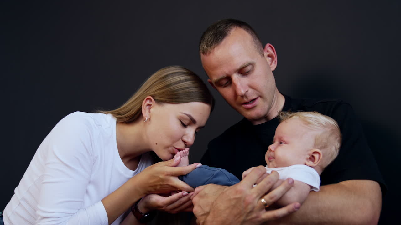 Man in black t-shirt holding his lovely infant son. Happy mom kisses her child's tiny foot. Black backdrop.