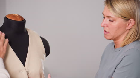 dress maker gently smooths beige waistcoat neckline on black mannequin while explaining fit details to colleague, assessing fabric drape, button alignment, seam precision inside minimalist studio