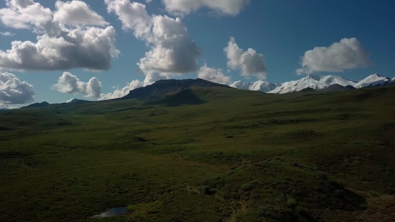 Splendido volo sulla natura selvaggia sopra le verdi alture e il ghiacciaio di Yukon Burwash verso la spettacolare catena montuosa dell'Anfiteatro coperta di neve sullo sfondo in una giornata di sole con cielo azzurro, Canada, avvicinamento aereo