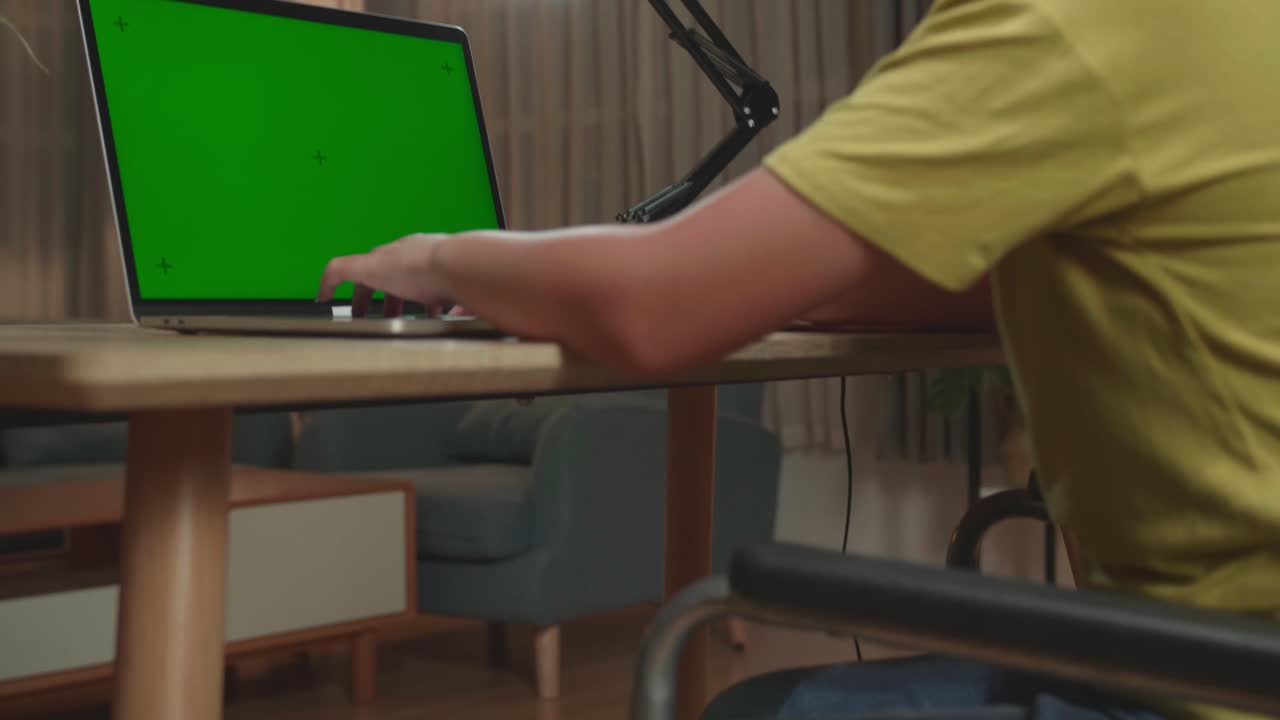 Back View Of Asian Kid Girl Sitting In A Wheelchair While Using Laptop Computer With Mock Up Green Screen At Home