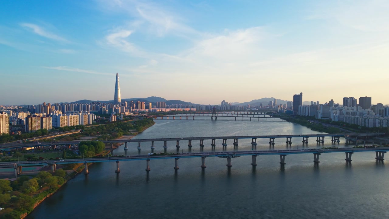 Una lenta ripresa aerea in parallasse cattura lo skyline di Seul al tramonto dorato, con la Lotte World Tower, i ponti del fiume Han e il Parco Gwangnaru Hangang illuminati da una luce calda, Corea del Sud.