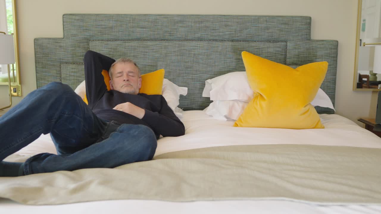 Man wearing clothes asleep on double bed with orange pillows, locked off