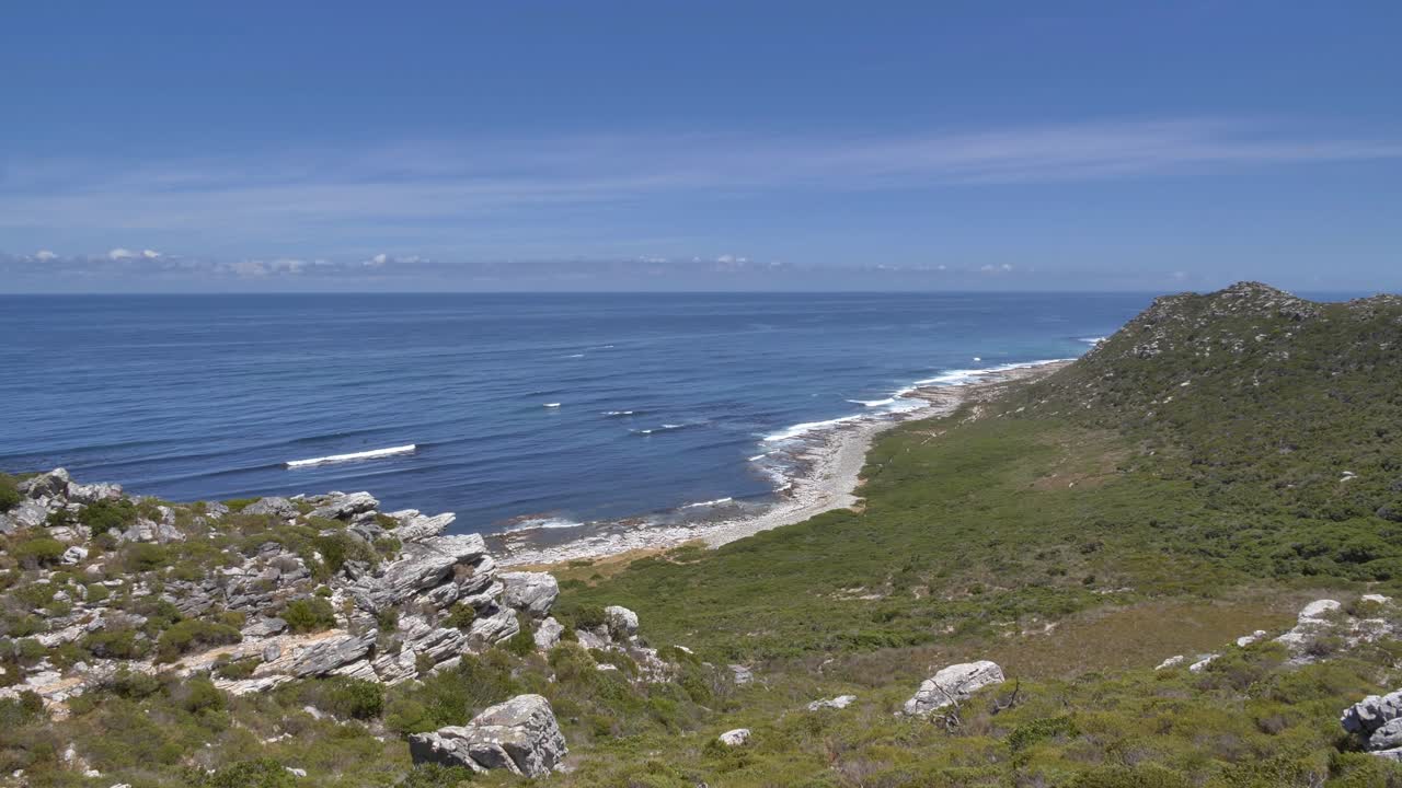 Ripresa con treppiede di un'ampia vista di una costa rocciosa con vegetazione verde lussureggiante che incontra l'oceano blu profondo sotto un cielo sereno al Capo di Buona Speranza, Riserva Naturale di Cape Point, Sudafrica