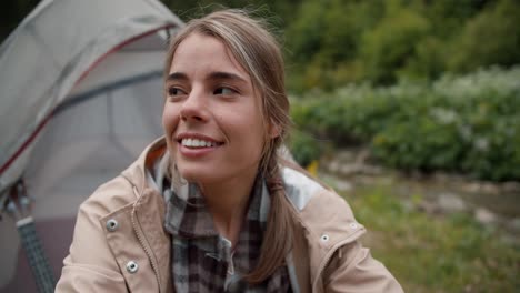 Portrait-of-a-happy-girl-blonde-tourist-who-sits-near-the-tent-on-the-background-of-the-forest