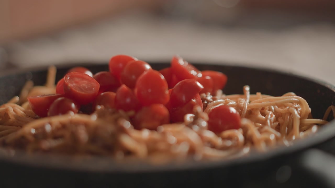 Vista dal basso di spaghetti cotti in una padella scura con pomodorini ciliegino tagliati a metà, salsa lucida, vapore leggero e consistenza fresca, creando un'atmosfera calda di cena casalinga con un leggero movimento dovuto alla recente preparazione.