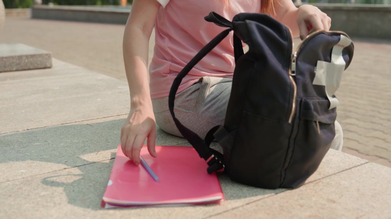 Young woman in pink shirt sits outdoors on stone steps opening her black backpack and taking out items including notebook with pen. scene features green trees distant buildings under bright