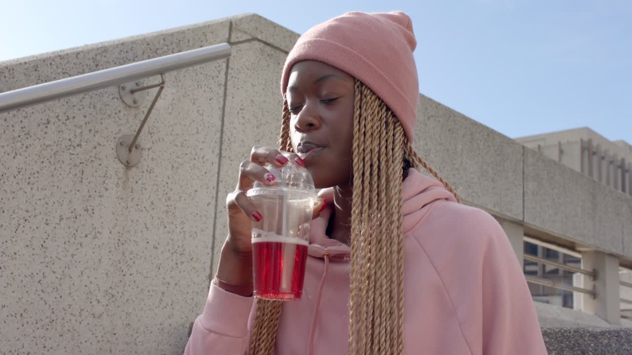 Afroamerikansk kvinde slukker tørsten og nipper til en rød drink med sugerør på trappen, med salat og plads til tekst.