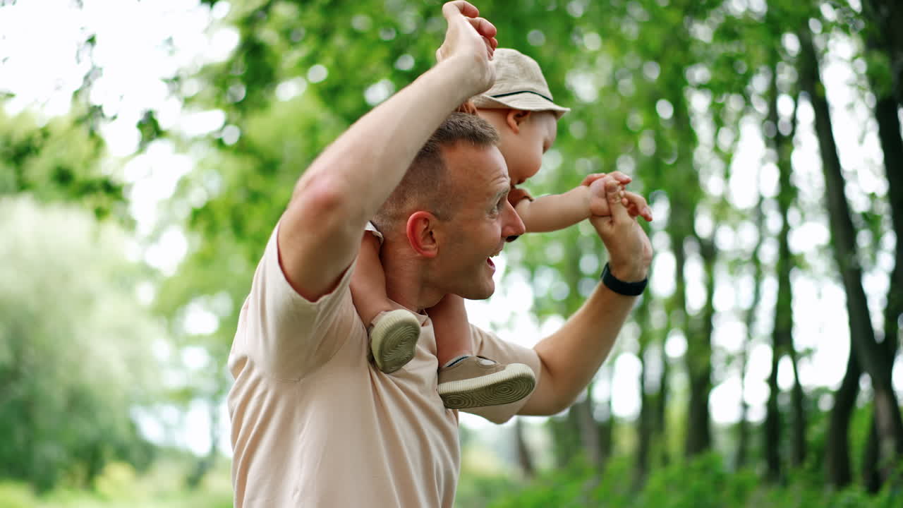 Caucasian father dances outdoors holding his son on the shoulders. Dad and baby having fun outdoors in summer.