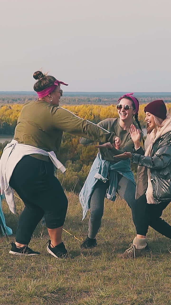 funny plump woman dances with girl friends by blue tent in camp on steep river bank in autumn evening slow motion