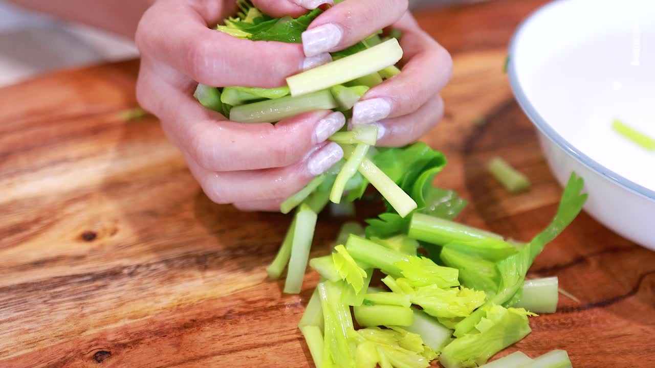 Hands arranging chopped celery on a wooden board and placing it into a bowl under bright lighting