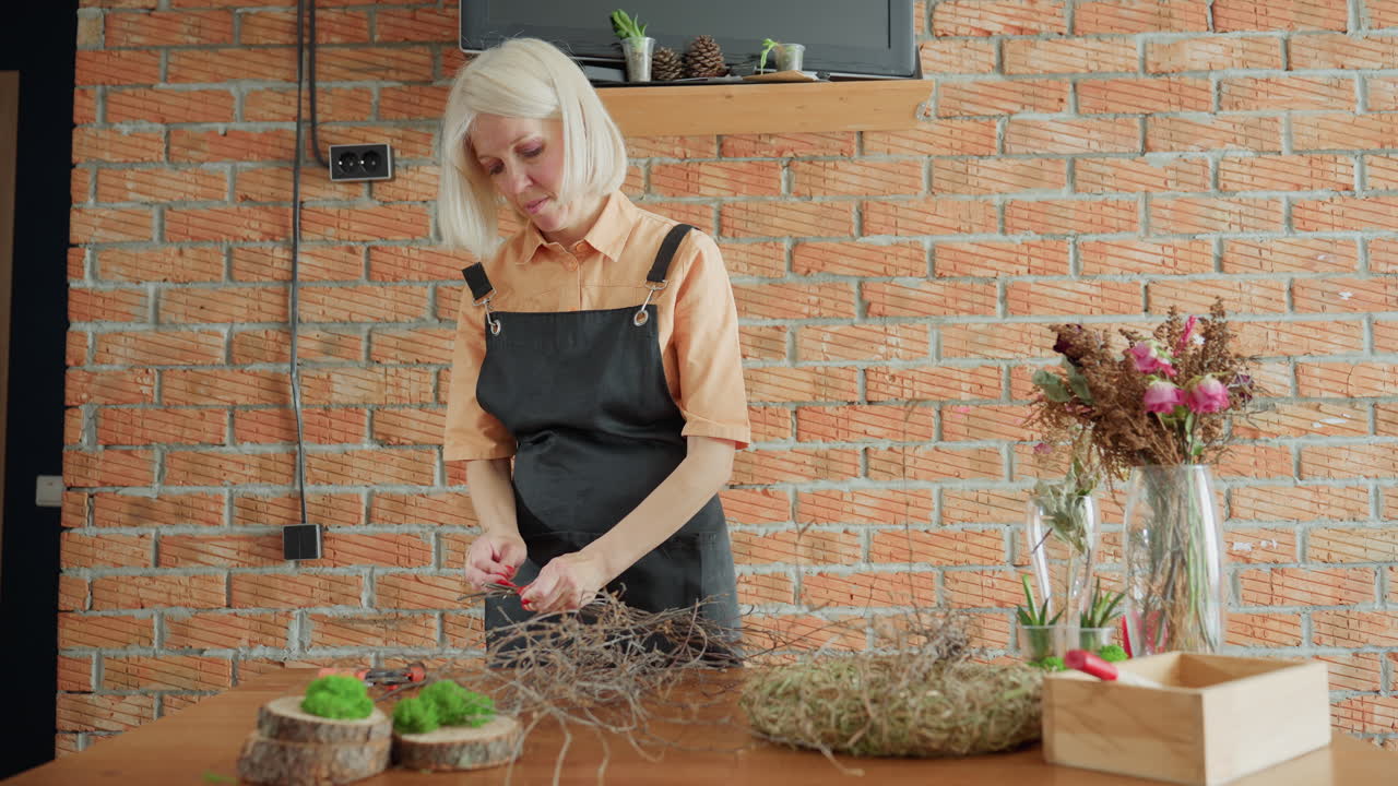 Woman in black apron working with dry twigs on wooden table arranging natural craft materials with moss and dried flowers in glass vase against brick wall background inside creative workshop space