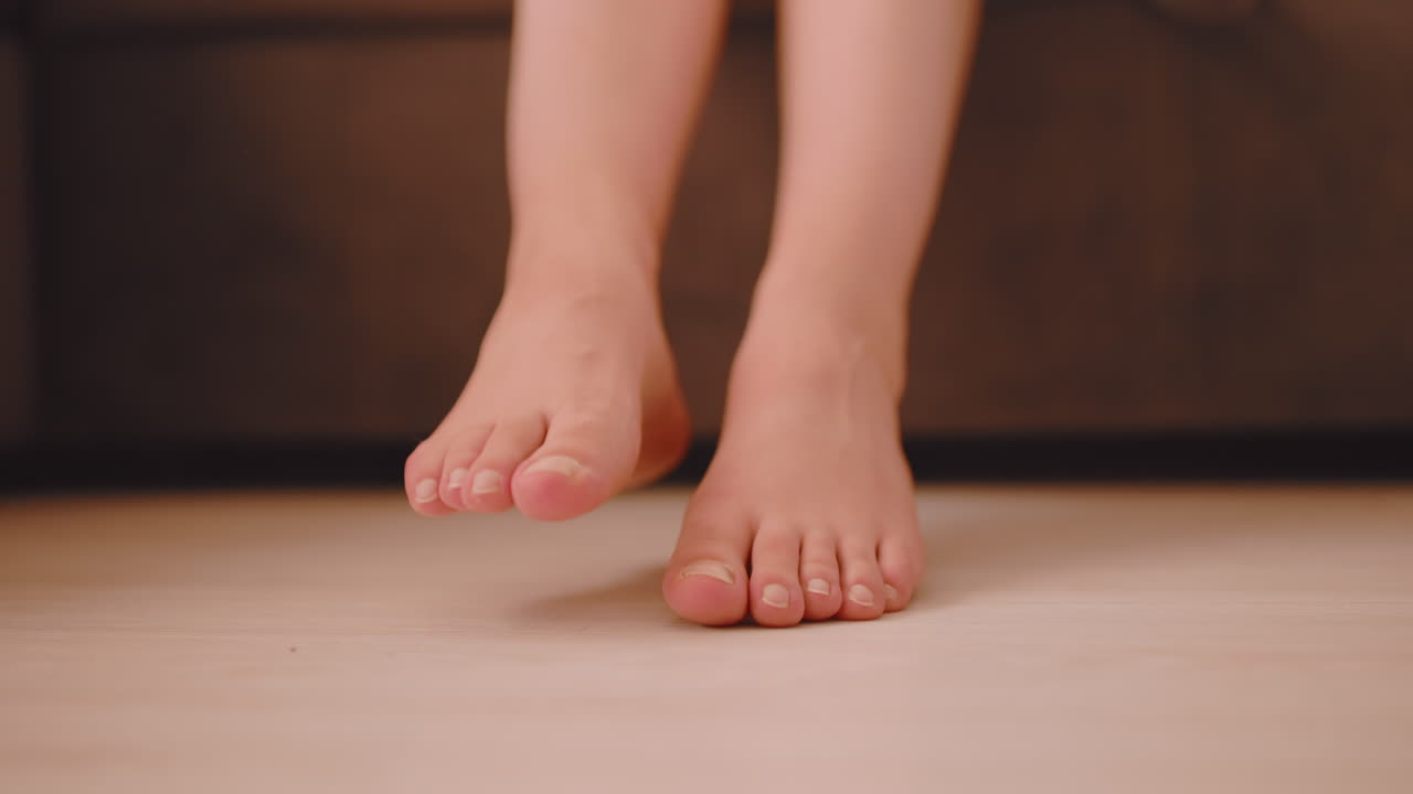 Close up of girl sitting on soft cushioned sofa gently wiggling bare feet just above smooth wooden floor with warm ambient lighting creating peaceful morning mood in quiet indoor environment
