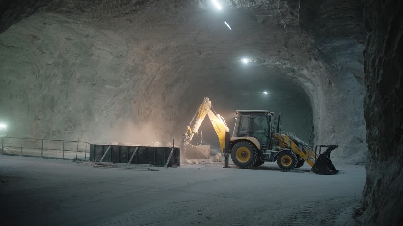 Static shot of a backhoe loader excavator working inside a vast, subterranean salt mine (salina). Dust fills the air under artificial lights, emphasizing the scale of the underground cavern