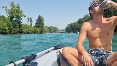 Attractive man relaxes on an inflatable boat tour along the turquoise Aare River in Bern, Switzerland, sipping a drink and enjoying the day amid lush riverside nature