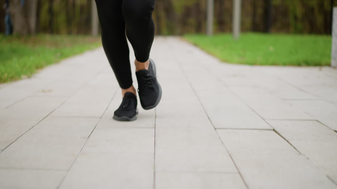 Close-up leg view of runner wearing black sneakers running on a paved track through park, blurred background of green grass, highlighting athletic movement and jogging activity