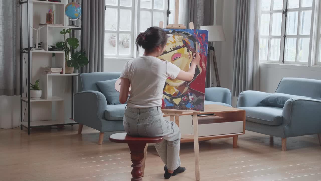 Hind View Of An Asian Artist Girl Sitting On Round Wooden Chair Without Backrest Holding Paintbrush Mixed Colour And Painting A Girl'S Face On The Canvas