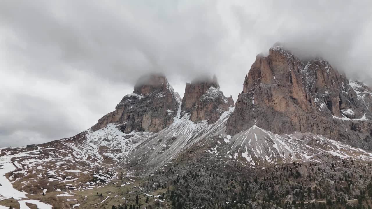 Flying backwards drone shot of the three iconic peaks of Sassolungo (Langkofel) in the Dolomites, Italy. Slowly pulling away, the mountains remain wrapped in clouds, revealing their towering presence