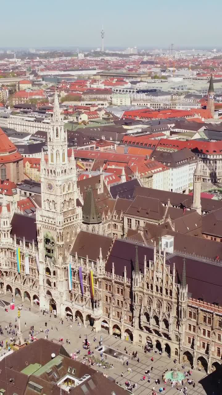Neues Rathaus at Marienplatz in Munich, Germany, revealing Gothic details, crowded square and urban rooftops with Olympiaturm visible on horizon under blue sky, vertical drone ascending and tilting