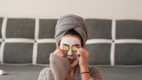 pretty black-haired woman with a towel on her head and a white cosmetic mask on the forms indulges in cucumber slices